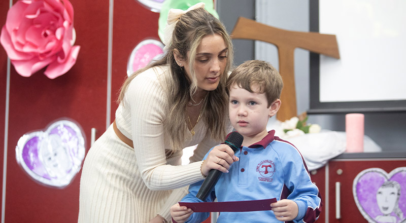A teacher holding a microphone to a students mouth while head reads at a Mother's Day event at St Bernadette's Primary School Dundas Valley
